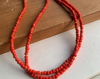 May include: A double-strand necklace featuring small, vibrant orange beads interspersed with tiny, metallic gold-toned spacer beads. The necklace is arranged in a circular loop, showcasing a consistent pattern of orange and gold elements throughout the design.