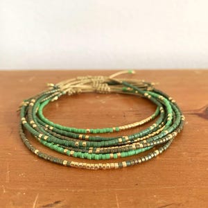 May include: A set of beaded bracelets in various shades of green and gold. The bracelets are made with small, colourful beads and are strung together on a neutral-coloured cord. The bracelets are arranged in a circular pattern.