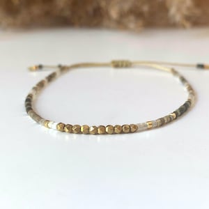 May include: Adjustable beaded bracelet with a mix of gold, white, and earth-toned beads. The bracelet has a sliding knot closure. The beads are small and faceted. The bracelet is displayed on a white surface, creating a delicate accessory.