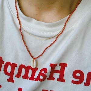 May include: A red beaded necklace with a small white seashell pendant. The necklace is layered over a gold chain. The necklace is worn by a person wearing a white t-shirt with the text "New York" and "Hamptons" printed in red.