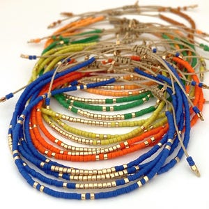 Puede incluir: Una colección de pulseras de cuentas coloridas. Las pulseras presentan cuentas azules, naranjas, verdes y amarillas, intercaladas con cuentas doradas. Están ensartadas en cordones de color natural.