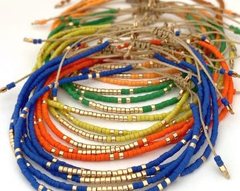 May include: A collection of adjustable bracelets made from colorful seed beads and metallic gold beads strung on tan cord. The bracelets feature vibrant blue, orange, green, and yellow beads arranged in patterns with gold accents and sliding knots.