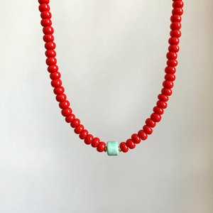 May include: A red beaded necklace featuring a light blue square bead at the centre. The necklace is composed of small, round red beads, strung together with gold-coloured accents. The necklace is set against a white backdrop.