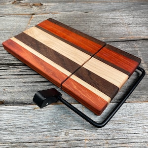 May include: A wooden cheese slicer with a multi-coloured striped design. The wood features alternating stripes of red, brown, and natural wood tones. It has a black metal wire and handle for slicing cheese. The slicer is approximately 25 cm long and 20 cm wide.