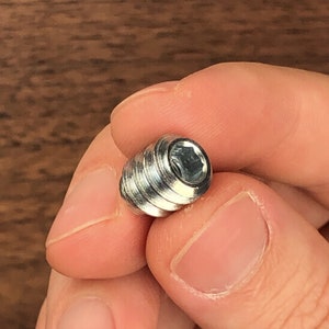 May include: Close-up of a silver metal set screw with a hex socket head. The screw has external threads and a cylindrical shape. The screw is held in a person's hand.