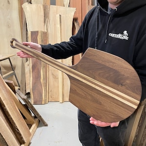 May include: A long-handled wooden pizza peel with a walnut wood surface and a light wood stripe down the centre. The peel has a rounded, paddle-shaped head and a curved handle.