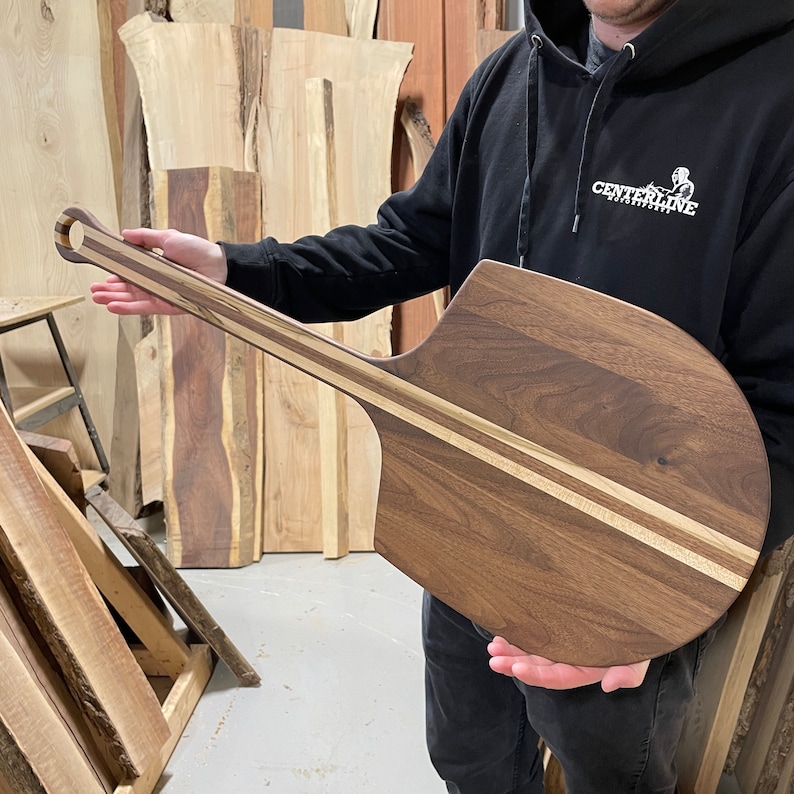 May include: A long-handled wooden pizza peel with a walnut wood surface and a light wood stripe down the centre. The peel has a rounded, paddle-shaped head and a curved handle.