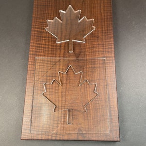 May include: A wooden plaque with a maple leaf cut-out design. The plaque has a dark brown wood grain finish. A clear acrylic maple leaf is placed on top, and another is set within a square cut-out.