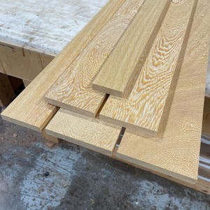 May include: A stack of unfinished, light-colored wooden planks with a prominent wood grain pattern. The planks vary in width and are arranged in a slightly overlapping manner, showcasing the natural texture and color variations of the wood.