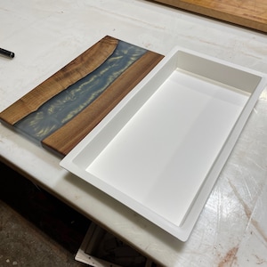 May include: A white rectangular mold for resin casting, next to a finished wooden cutting board with a blue and gold resin inlay.