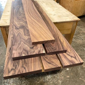 May include: A stack of rectangular walnut wood boards featuring prominent dark brown grain patterns and swirling wood textures. The boards are arranged in a staggered formation, showcasing the natural variations in the wood's colour and surface.