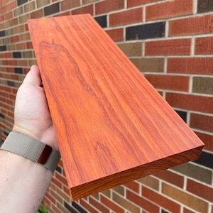 May include: A close-up of a long, rectangular piece of reddish-brown wood with a smooth, polished surface. The wood has a natural grain pattern and is held in a hand.