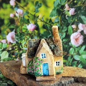 Könnte beinhalten: Eine Miniatur-Cottage-Figur mit einer blauen Tür, kleinen Fenstern und einem Schornstein. Das Cottage besteht aus beigen und braunen Materialien und ist mit einem Kletterrosenbusch verziert. Die Figur steht auf einem braunen Stein, mit einem Hintergrund aus rosa Rosen und grünem Laub.