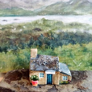 Pode incluir: Uma miniatura de casa com telhado cinzento texturizado, paredes bege, uma porta azul e uma pequena planta em vaso perto da entrada. A estrutura tem duas janelas visíveis com molduras brancas e assenta sobre uma pedra escura e irregular.