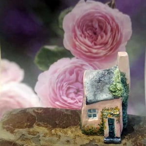 Puede incluir: Una figura de casa de campo en miniatura pintada a mano. La casa rosa tiene un tejado gris, una puerta azul y una pequeña ventana. El follaje verde adorna la puerta y el tejado. El fondo presenta suaves rosas rosas.
