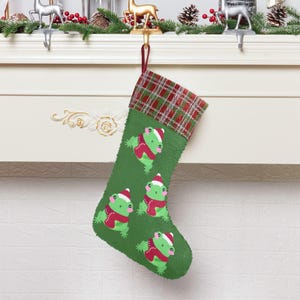 May include: Green Christmas stocking with a red and green plaid cuff. The stocking features a repeating pattern of green frogs wearing red scarves.