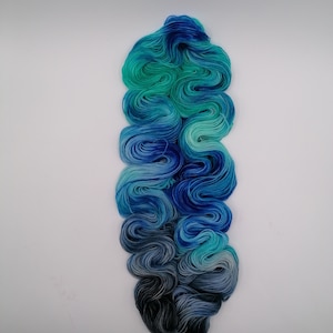 May include: A skein of yarn with a gradient of blue, teal, and black colors. The yarn is wound into a loose coil.