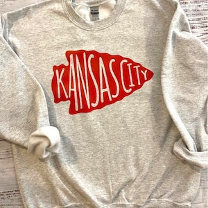 May include: A gray sweatshirt with a red arrow design that says "Kansas City" in white lettering.