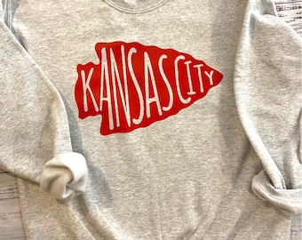 Può includere: Una felpa grigia con un design a freccia rossa che dice "Kansas City" in lettere bianche.