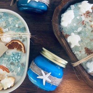 May include: Three blue glass candle jars with lids, two are round and one is square. The square jar has a white label that says "Esmay Melts Coastal". The round jars are decorated with twine and a white starfish. The square jar is filled with white wax and blue glass pieces. The round jars are filled with blue wax.