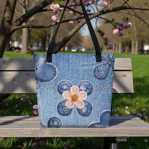 May include: Denim tote bag with black handles. The bag features a patchwork design with blue denim patches and a large, embroidered flower in pink and yellow. The bag is sitting on a wooden bench in a park setting.