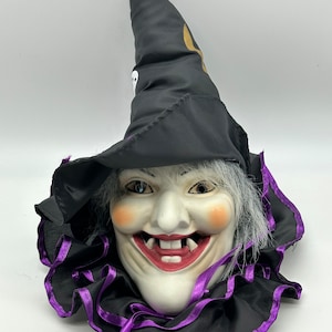 May include: A Halloween witch mask with a black pointed hat featuring a gold crescent moon and a white skull. The mask has a smiling face with rosy cheeks, grey hair, and a black collar trimmed with purple ribbon.