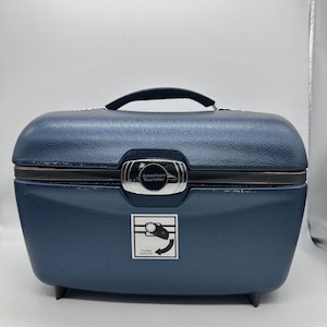 May include: A navy blue American Tourister vanity case with a black handle and silver clasp. The case has a textured surface and a graphic with the words "Turn Down".