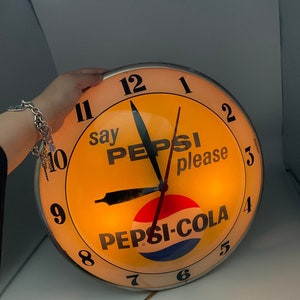May include: A vintage Pepsi-Cola clock with a yellow face and black hands. The clock has a red, white, and blue Pepsi-Cola logo in the center. The clock face also says "say Pepsi please".