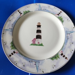 May include: White ceramic dinner plate with a blue and white lighthouse and a small pink house on a blue and white sea background. The plate has a blue rim and a white border with a repeating pattern of seagulls and sailboats.