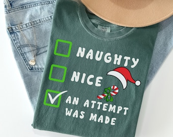 May include: A green t-shirt with the text "NAUGHTY NICE AN ATTEMPT WAS MADE" in white. The shirt features a Santa hat and candy cane graphic. Light blue jeans and a tan hat are also in the image.