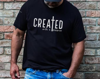 Puede incluir: Camiseta negra con texto blanco que dice "CREATED with a PURPOSE" y un gráfico de cruz blanca.
