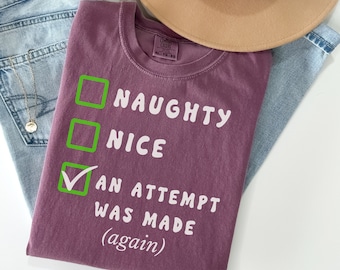 May include: A purple t-shirt with a white check list that says "Naughty", "Nice", and "An attempt was made (again)". The check box next to "An attempt was made (again)" is checked.