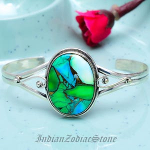 Op de afbeelding: Zilveren manchetarmband met een ovale, veelkleurige steen met groene, blauwe en witte tinten. De steen is gezet in een zilveren bezel, en de armband heeft een open ontwerp met decoratieve krullen. De tekst "Indian Zodiac Stone" staat onderaan.