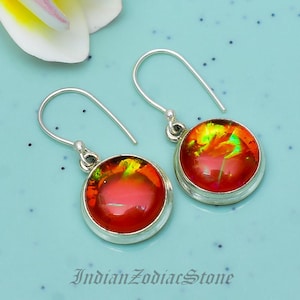 May include: A pair of drop earrings featuring round, vibrant red cabochons with iridescent green and yellow flecks set in silver-toned metal. The earrings have curved silver-toned ear wires and are displayed with the text IndianZodiacStone.