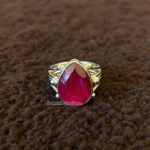 Puede incluir: Un anillo de plata con una gran piedra preciosa roja en forma de lágrima. El anillo tiene una banda adornada con pequeños adornos redondos. El texto "IndianZodiacStone" es visible en el anillo.