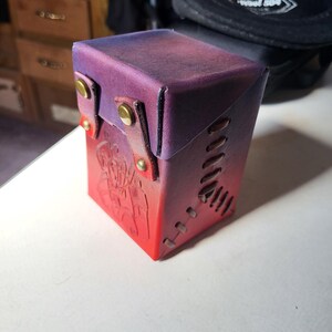 May include: A rectangular leather deck box featuring a gradient color scheme that transitions from deep purple at the top to vibrant red at the base. The box includes a flap secured by two brass-colored metal snaps and decorative side lacing.