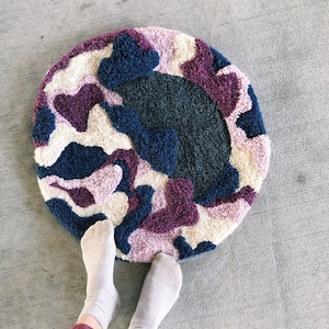 May include: A round, colorful rug with a pink, purple, blue, and white abstract pattern. The rug is made of yarn and has a fluffy texture.