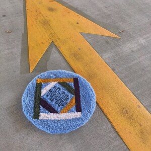 May include: A blue round rug with a geometric pattern in shades of yellow, green, purple, and white. The rug is on a gray concrete surface with a yellow arrow painted on it.