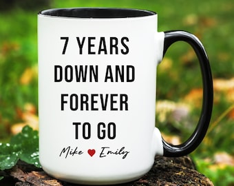 May include: A white ceramic mug with a black interior and handle. The mug features the text "7 YEARS DOWN AND FOREVER TO GO" in bold black letters, with "Mike ❤️ Emily" below. The mug is a gift for a couple.