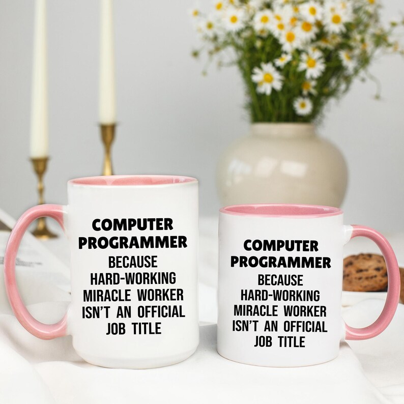 May include: Two white coffee mugs with pink handles and rims. The mugs have the text "COMPUTER PROGRAMMER BECAUSE HARD-WORKING MIRACLE WORKER ISN'T AN OFFICIAL JOB TITLE" in black. A vase of flowers is in the background.