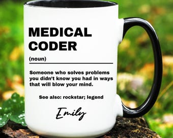 May include: White ceramic mug with a black handle and rim. The mug features the text "MEDICAL CODER" above a definition, and the name "Emily" in script. The mug is sitting on a wooden surface.