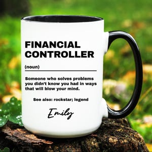 May include: White ceramic mug with a black handle and rim. The mug features the text "FINANCIAL CONTROLLER" in bold black letters, along with a definition and the name "Emily" in script. The mug is sitting on a wooden surface.