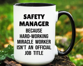 May include: White ceramic mug with a black interior and handle. The mug features the text "SAFETY MANAGER" above the phrase "BECAUSE HARD-WORKING MIRACLE WORKER ISN'T AN OFFICIAL JOB TITLE" in bold black letters.