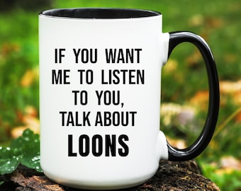 May include: White ceramic mug with a black interior, handle, and rim. The mug features the text "IF YOU WANT ME TO LISTEN TO YOU, TALK ABOUT LOONS" in black, bold font. The mug is set against a blurred green background.