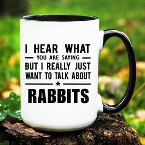 May include: White ceramic mug with a black handle and rim. The mug features the text "I HEAR WHAT - YOU ARE SAYING - BUT I REALLY JUST WANT TO TALK ABOUT RABBITS" in black, with a star graphic.
