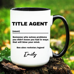 May include: White ceramic mug with a black interior and handle. The mug features the text "TITLE AGENT" in bold black letters, along with a definition and the name "Emily" in script. The mug is sitting on a wooden surface.