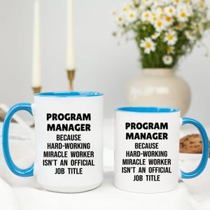 May include: Two white ceramic mugs with blue interiors and handles. The mugs have the text "PROGRAM MANAGER BECAUSE HARD-WORKING MIRACLE WORKER ISN'T AN OFFICIAL JOB TITLE" in black. A vase of flowers is in the background.