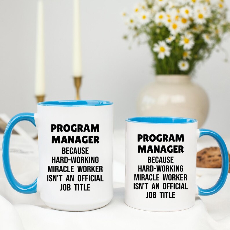 May include: Two white ceramic mugs with blue interiors and handles. The mugs have the text "PROGRAM MANAGER BECAUSE HARD-WORKING MIRACLE WORKER ISN'T AN OFFICIAL JOB TITLE" in black. A vase of flowers is in the background.