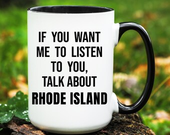 May include: White ceramic mug with a black interior and handle. The mug features the text: "IF YOU WANT ME TO LISTEN TO YOU, TALK ABOUT RHODE ISLAND" in a bold, black font. The mug is set against a blurred green background.