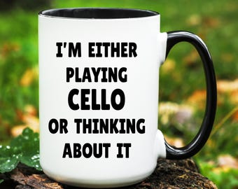 May include: White ceramic mug with a black interior and handle. The mug features the text "I'M EITHER PLAYING CELLO OR THINKING ABOUT IT" in bold black letters. The mug is sitting on a wooden surface.